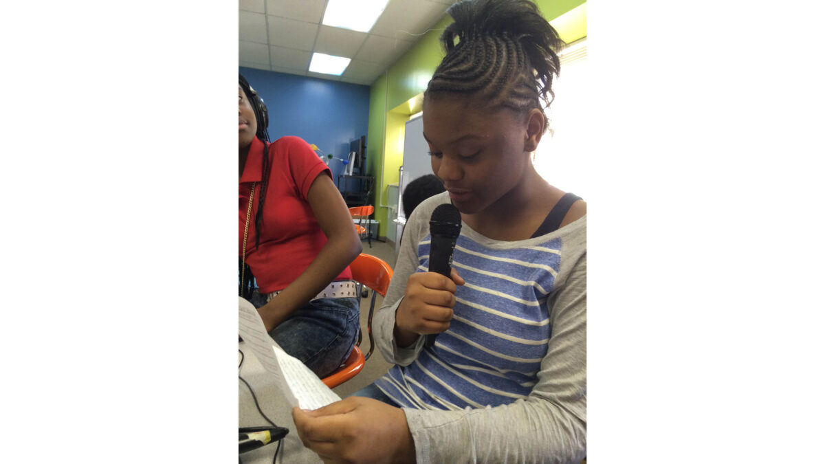 A youth looking at another youth holding a piece of paper and speaking into a microphone while sitting in a classroom