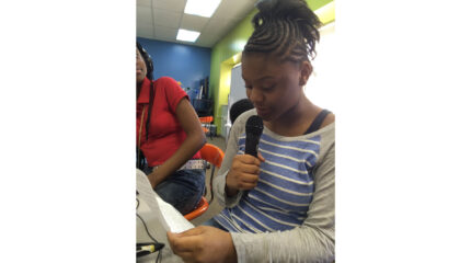 A youth looking at another youth holding a piece of paper and speaking into a microphone while sitting in a classroom