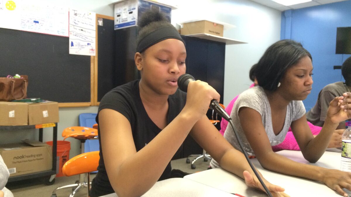 Two youth sitting at a white table in a classroom, one holding and speaking into a microphone