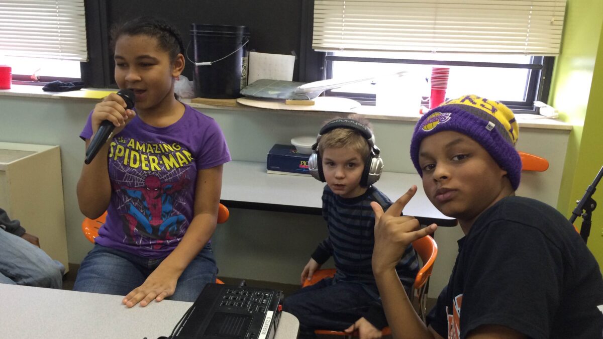 A youth holding and speaking into a microphone, a youth wearing headphones sitting, and another youth sitting and making a rock sign with their hands, all in a classroom.
