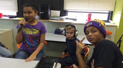 A youth holding and speaking into a microphone, a youth wearing headphones sitting, and another youth sitting and making a rock sign with their hands, all in a classroom.