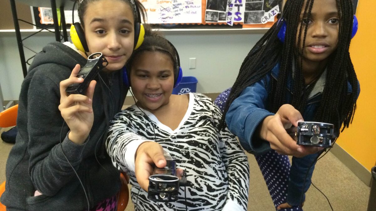 Three youth wearing headphones holding portable recorder up to the camera in a classroom