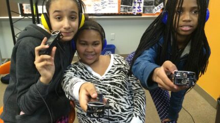 Three youth wearing headphones holding portable recorder up to the camera in a classroom
