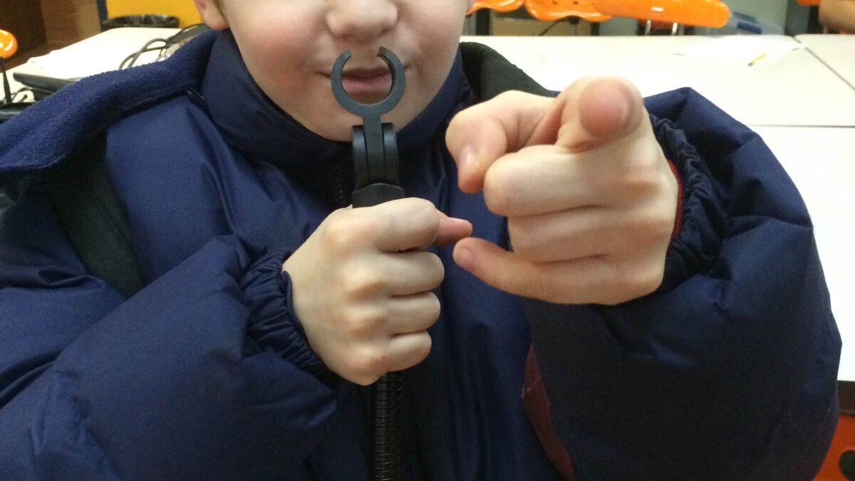 A youth holding a small microphone stand to their lips and pointing at the camera in a classroom