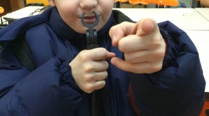 A youth holding a small microphone stand to their lips and pointing at the camera in a classroom