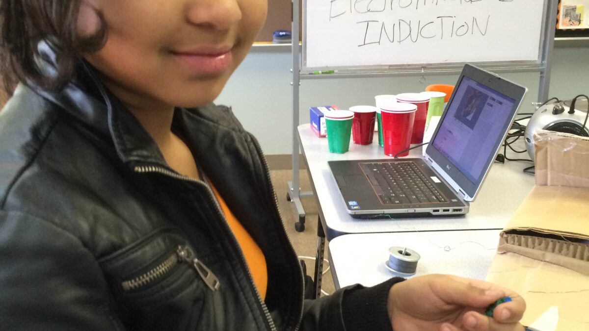 A youth interacting with copper wiring sitting next to a laptop computer and a whiteboard in a classroom