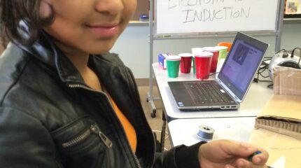 A youth interacting with copper wiring sitting next to a laptop computer and a whiteboard in a classroom