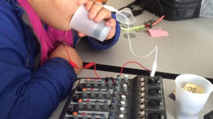 A youth pretending to drink from a cup with wires connected to a mixer in a classroom
