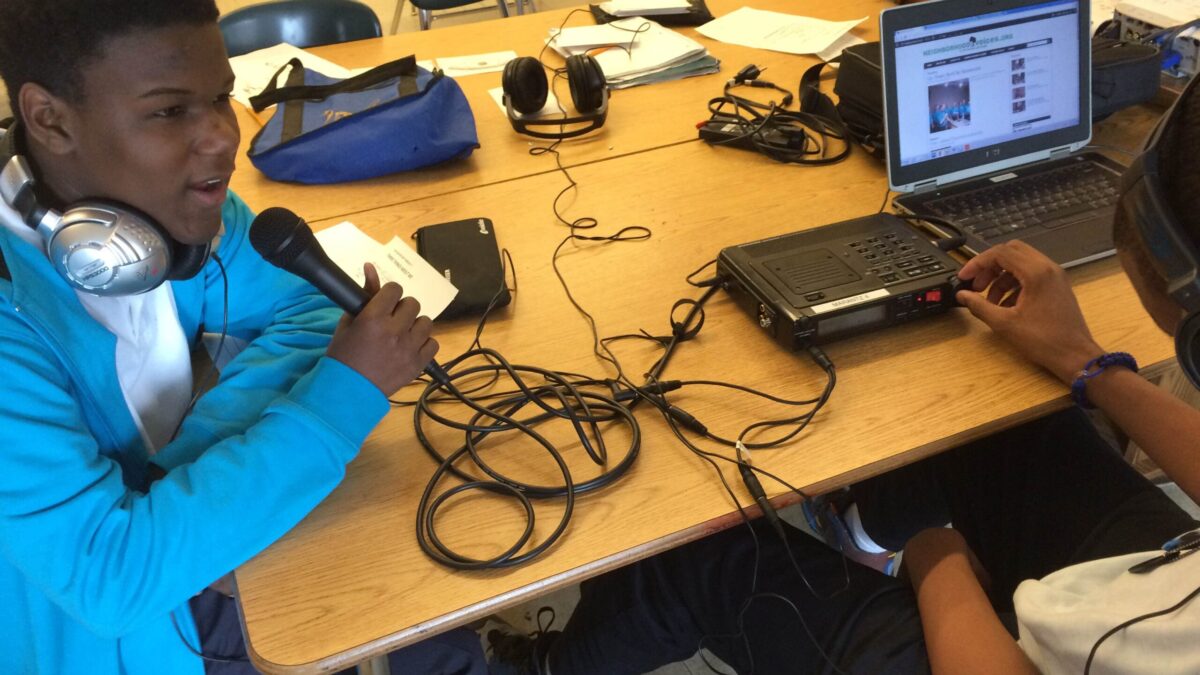 A youth wearing headphones speaking into a microphone connected to a laptop computer on a classroom desk