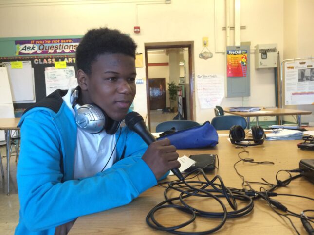 A youth wearing headphones holding and speaking into a microphone sitting at a classroom desk