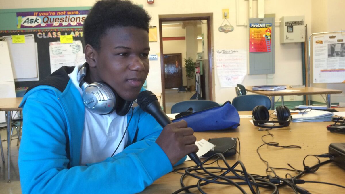 A youth wearing headphones holding and speaking into a microphone sitting at a classroom desk