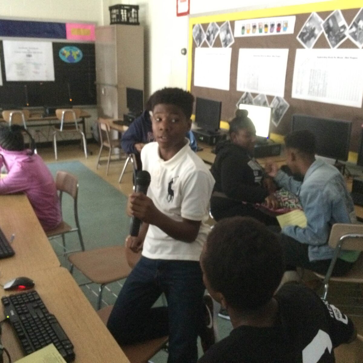 IMG_1059 Five youth sitting at computers on classroom desks and a youth holding a microphone sitting on their knees on a chair