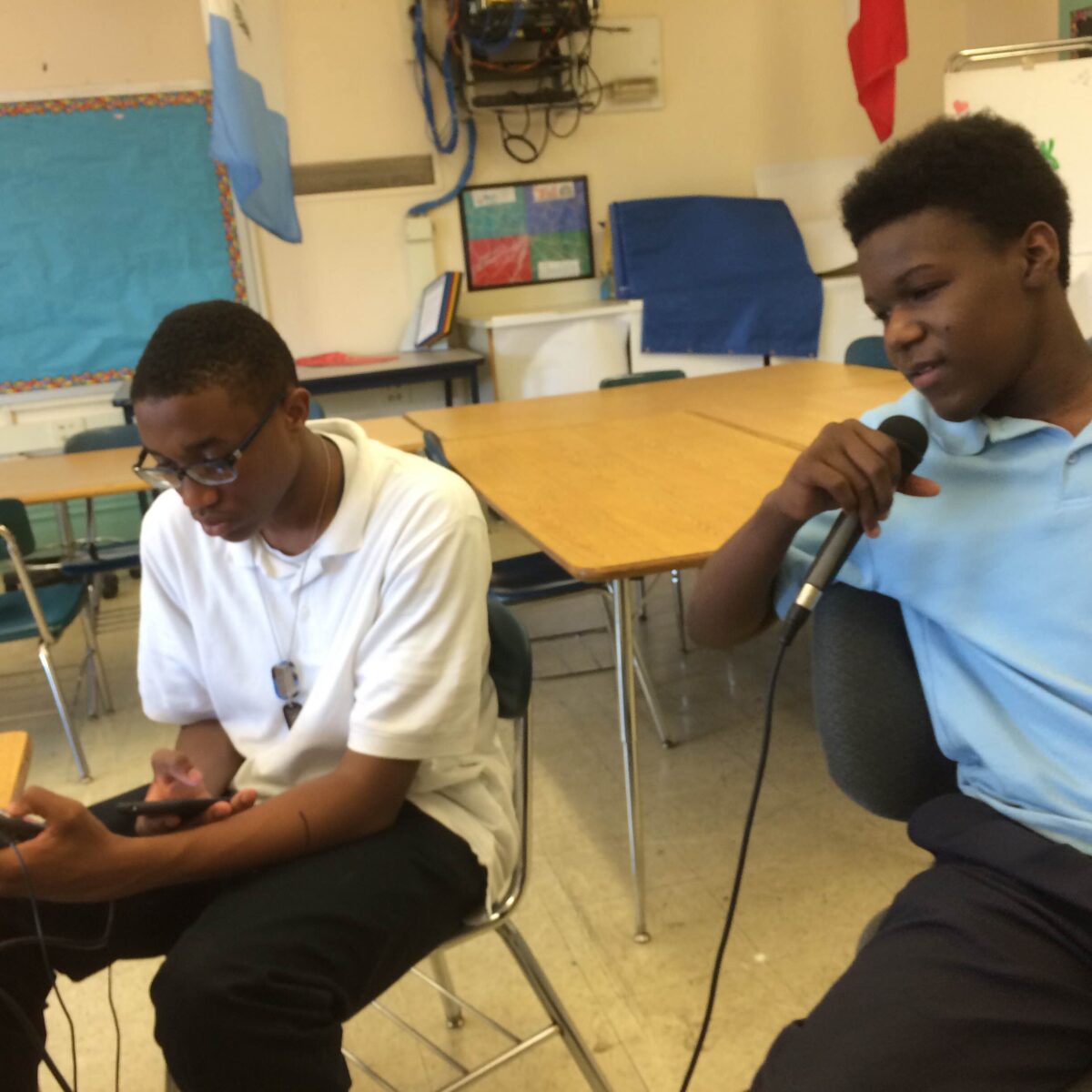 Two teens sitting in a classroom, one holding and speaking into a microphone, the other holding a portable microphone and looking at their smartphone