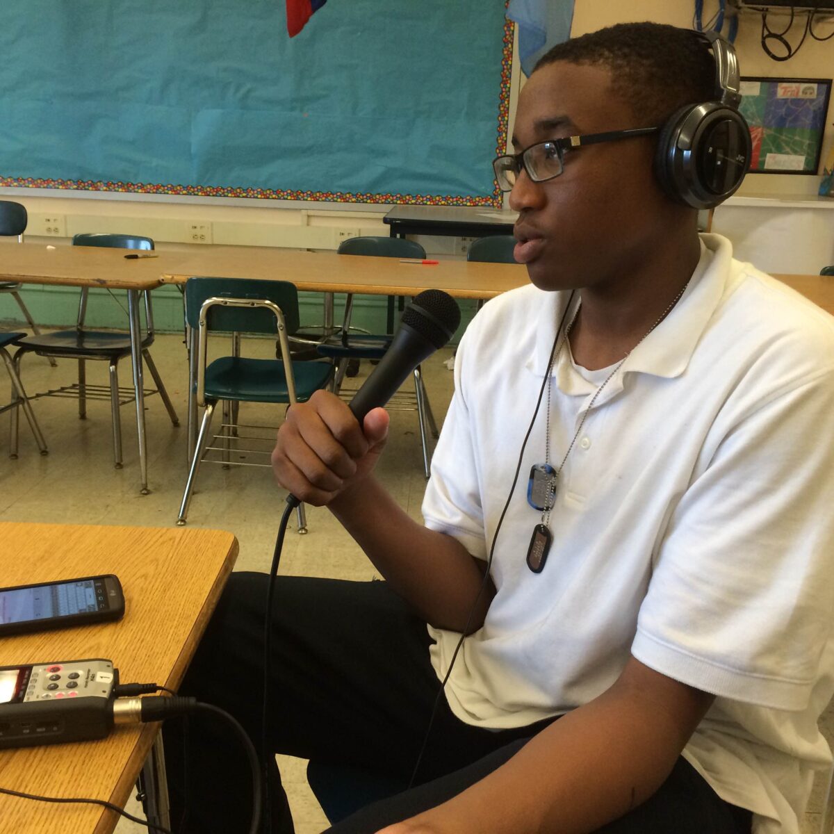 A teen wearing headphones holding and speaking into a microphone connected to a portable recorder on a classroom desk