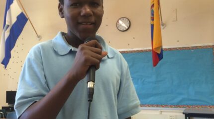 A teen holding and speaking into a microphone standing in a classroom