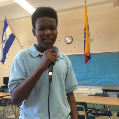 A teen holding and speaking into a microphone standing in a classroom