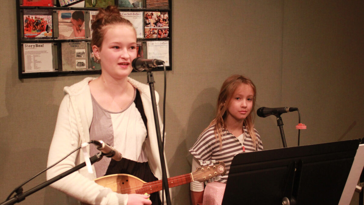 A youth holding a tamburitza and speaking into a microphone and a youth standing behind a microphone in a radio studio