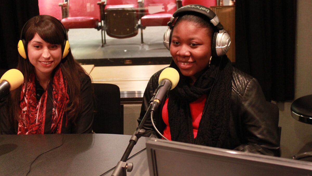 A teen and an adult wearing headphones speaking into microphones in a radio studio