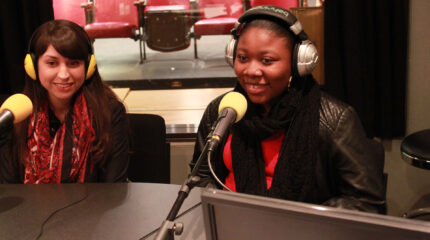 A teen and an adult wearing headphones speaking into microphones in a radio studio