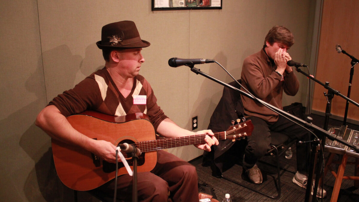 An adult playing an acoustic guitar and singing into a microphone and an adult playing a harmonica into a microphone into a microphone in a radio studio