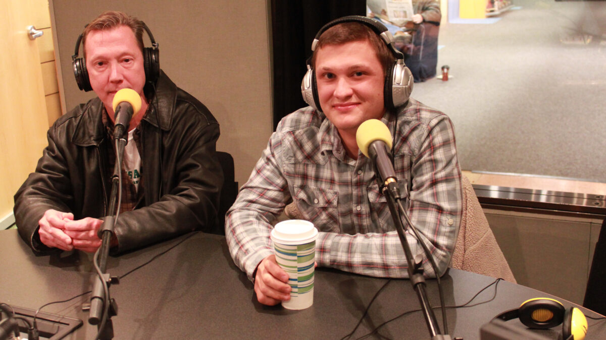 Two adults wearing headphones sitting behind microphones in a radio studio
