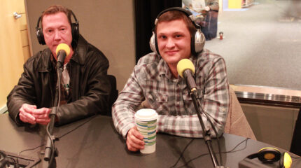 Two adults wearing headphones sitting behind microphones in a radio studio