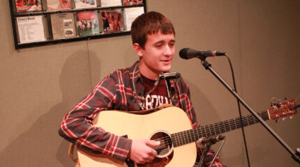 A teen holding an acoustic guitar and speaking into a microphone in a radio studio