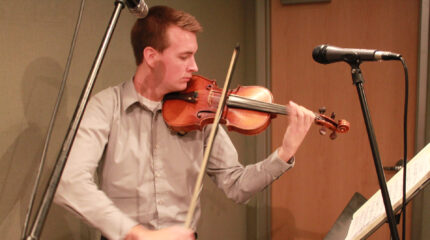 A teen playing a violin looking at sheet music on a music stand in a radio studio