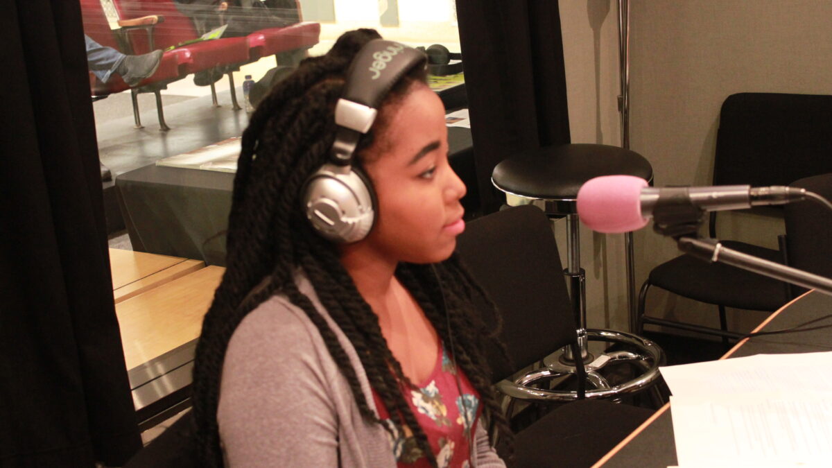 A teen wearing headphones sitting behind a microphone in a radio studio