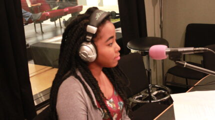 A teen wearing headphones sitting behind a microphone in a radio studio