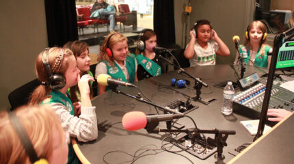 Seven youth wearing headphones speaking into microphones in a radio studio