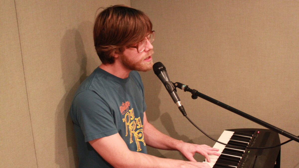 An adult playing piano keyboard and singing into a microphone in a radio studio