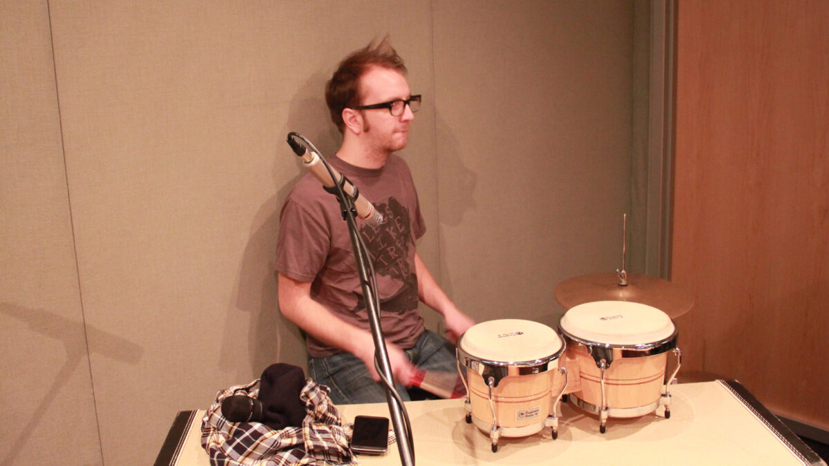 An adult playing a drum set sitting behind bongos in a radio studio