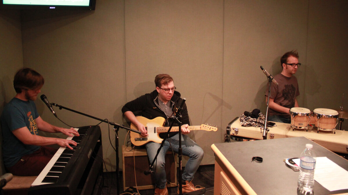 An adult playing a piano keyboard, an adult playing an electric guitar, and an adult playing a drum set in a radio studio