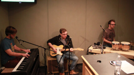 An adult playing a piano keyboard, an adult playing an electric guitar, and an adult playing a drum set in a radio studio