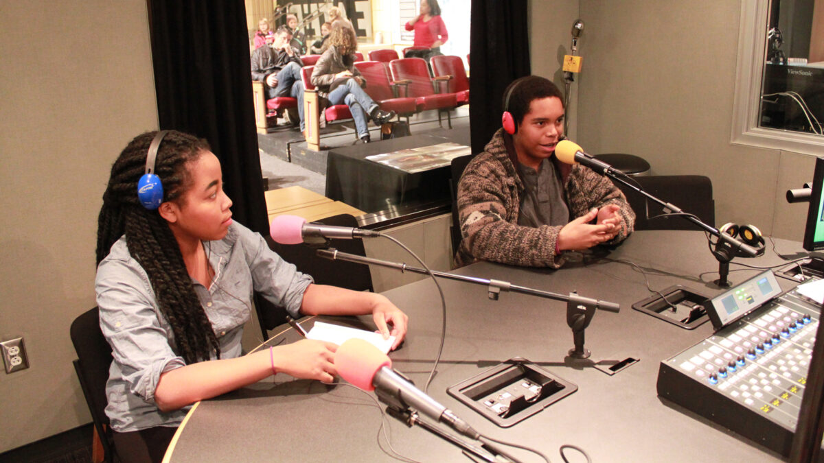 Two teens wearing headphones speaking into microphones in a radio studio