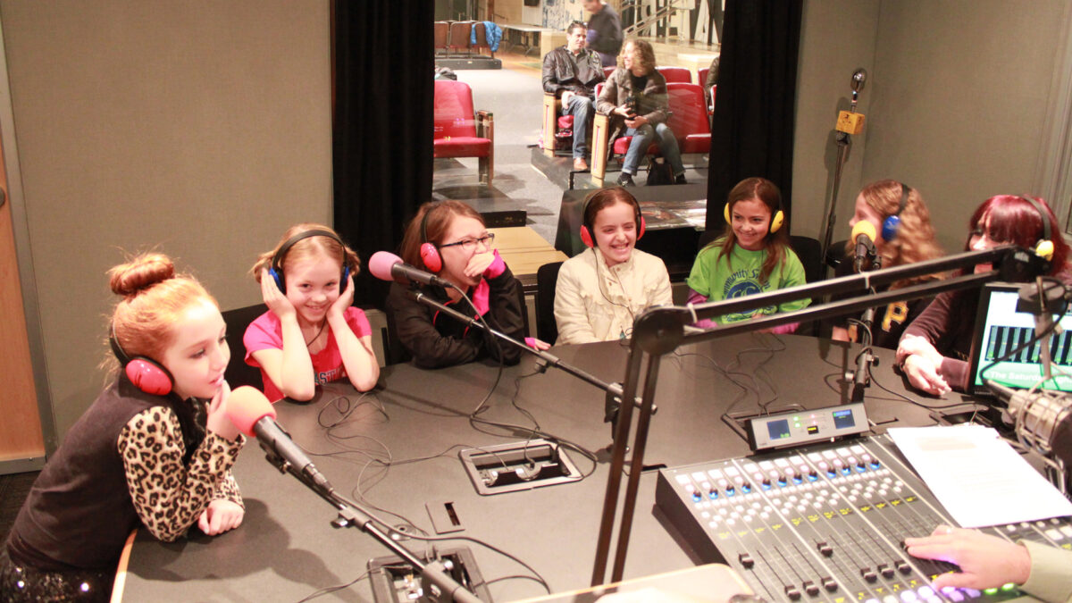 Six youth and an adult wearing headphones speaking into microphones in a radio studio