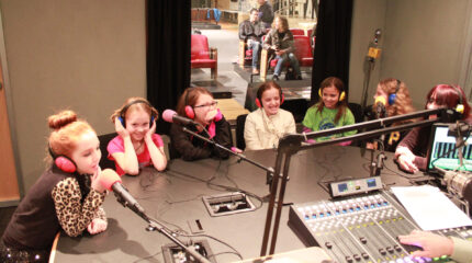 Six youth and an adult wearing headphones speaking into microphones in a radio studio