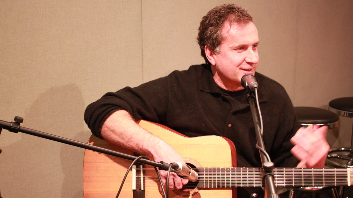 An adult playing acoustic guitar and singing into a microphone in a radio studio
