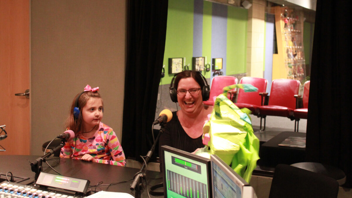A youth wearing headphones looking at an adult wearing headphones smiling while holding a green papier mache puppet in a radio studio