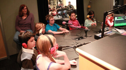 Six youth wearing headphones sitting behind microphones and an adult sitting in the background in a radio studio
