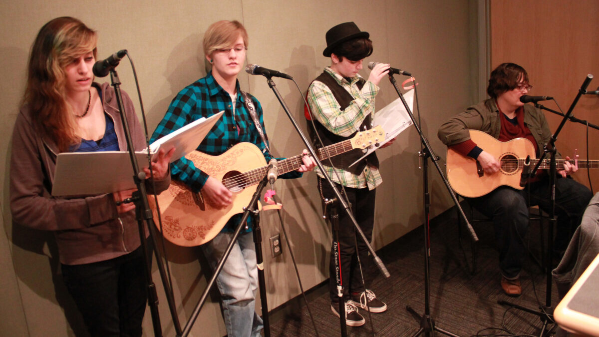 Four teens singing into microphones, two holding binders, two holding guitars, in a radio studio