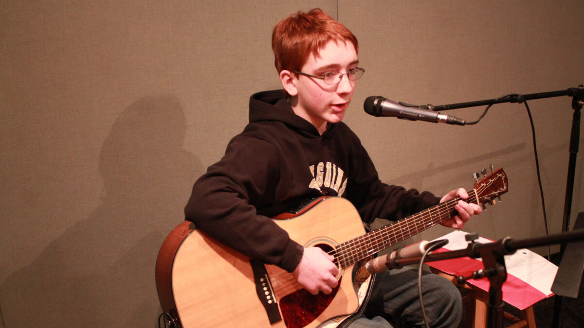 A youth playing an acoustic guitar and singing into a microphone in a radio studio