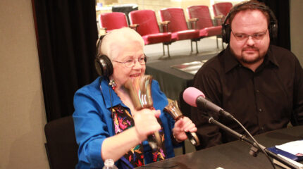 Two adults wearing headphones, one ringing two handbells, in a radio studio