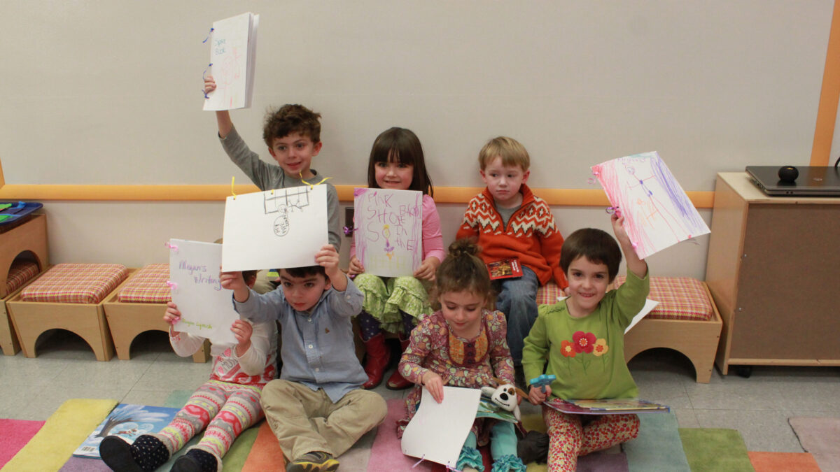 Seven children holding up drawings while sitting on a multicolored floor