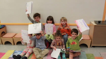 Seven children holding up drawings while sitting on a multicolored floor