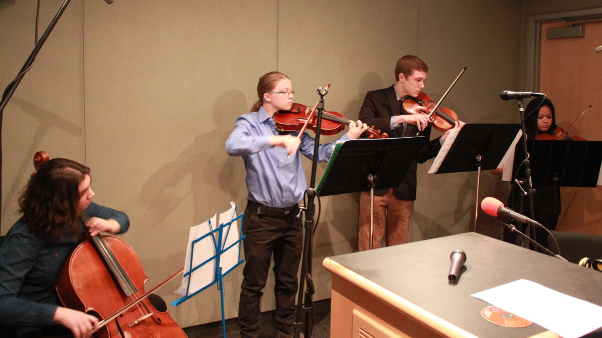 A teen playing cello, a teen playing viola, and two teens playing violins, all looking at sheet music on music stands in a radio studio