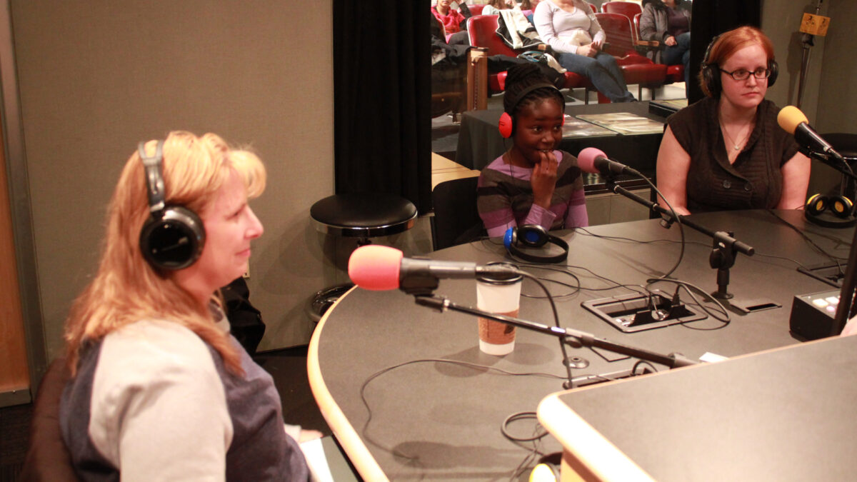 Two adults and a youth wearing headphones speaking into microphones in a radio studio