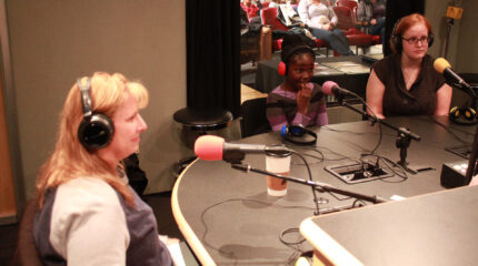 Two adults and a youth wearing headphones speaking into microphones in a radio studio