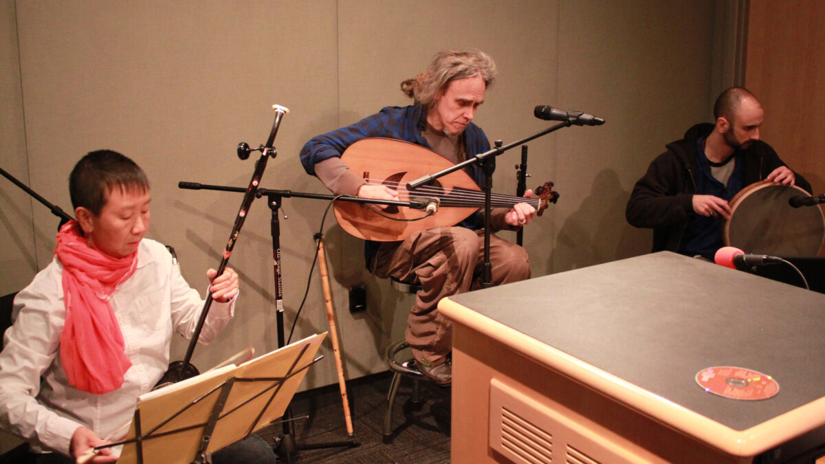 An adult playing an oriental string instrument, an adult playing an ethnic string instrument, and an adult playing a percussion instrument in a radio studio